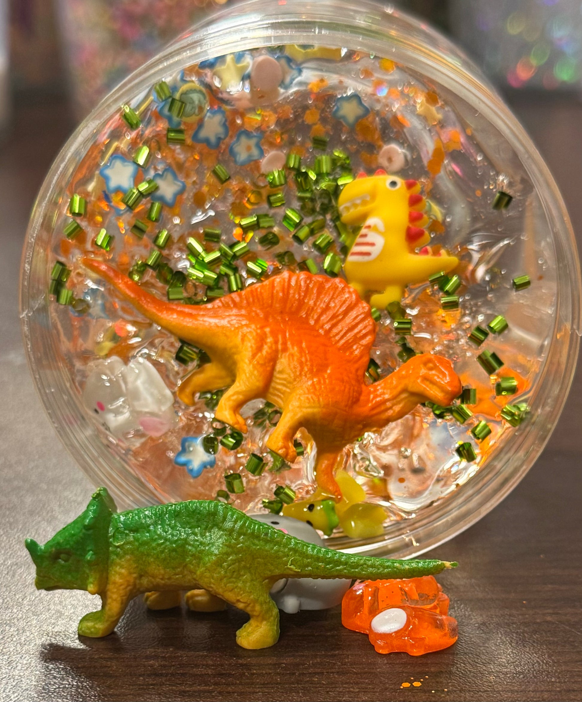 Dinosaur toys in a clear container with colorful contents on a wooden surface