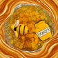 Jar filled with honeycomb-like substance, a toy bee, and a 'Honey' sign held by a person.